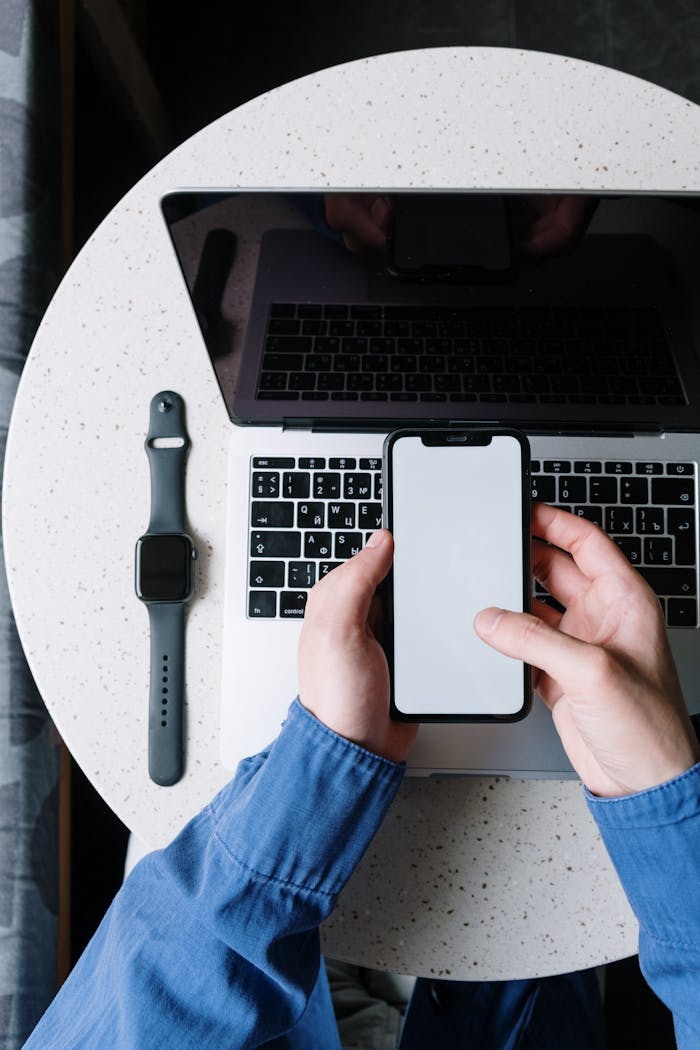 Top view of a person using a smartphone above a laptop with a smartwatch on the table.
